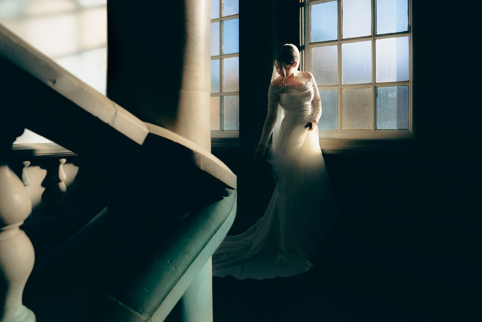 Bride in lace gown standing at a tall window, side-lit by warm afternoon sun
