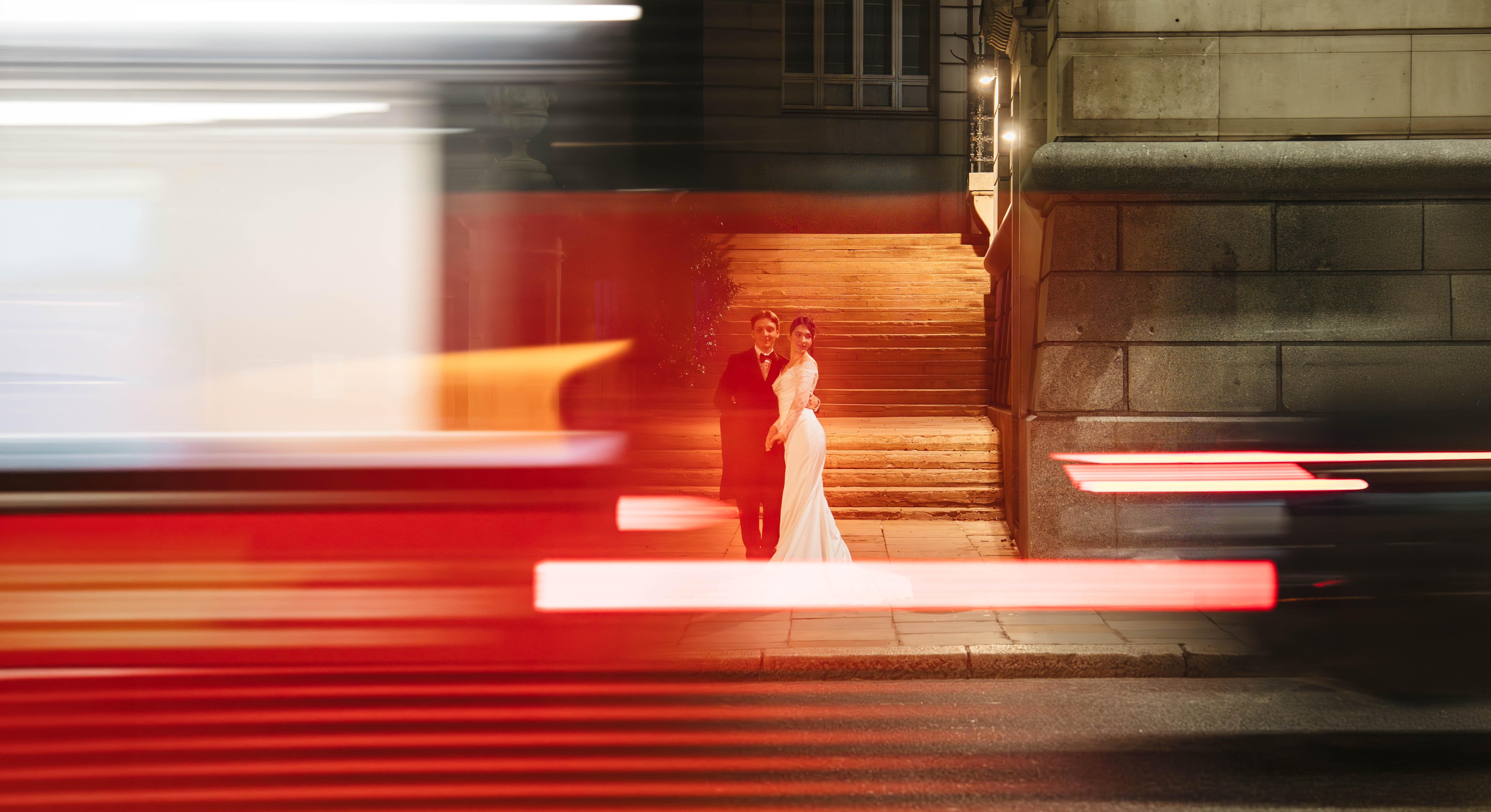 A couple holds still on lit stone steps as the streaks of a passing London bus blur across the foreground